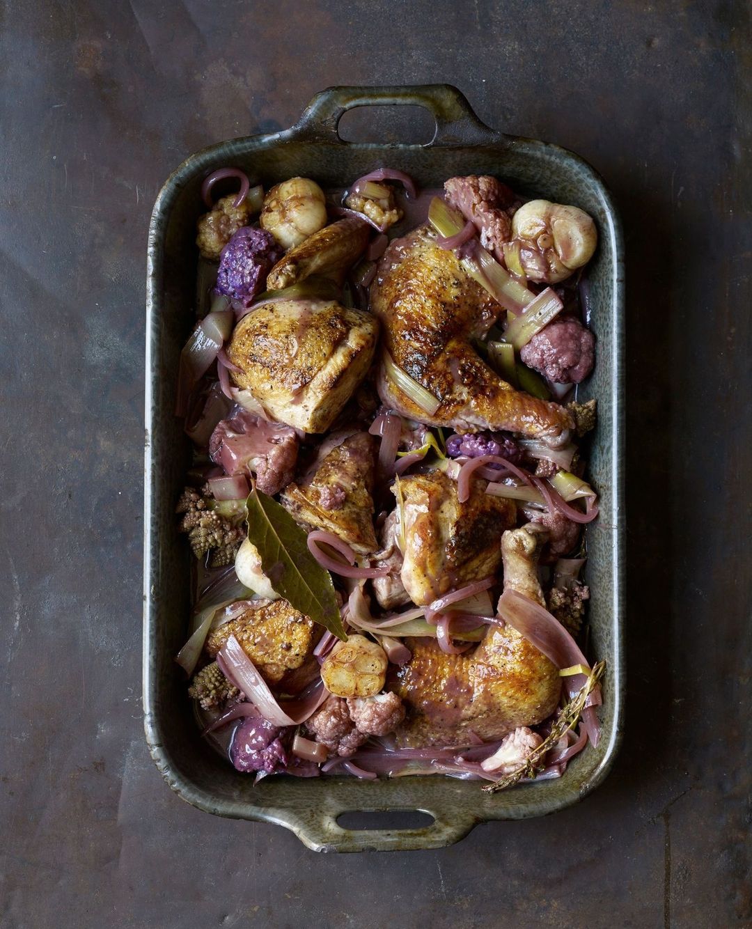 Overhead shot of roasted chicken pieces with vegetables, including onions and cauliflower, arranged in a dark metal baking pan.