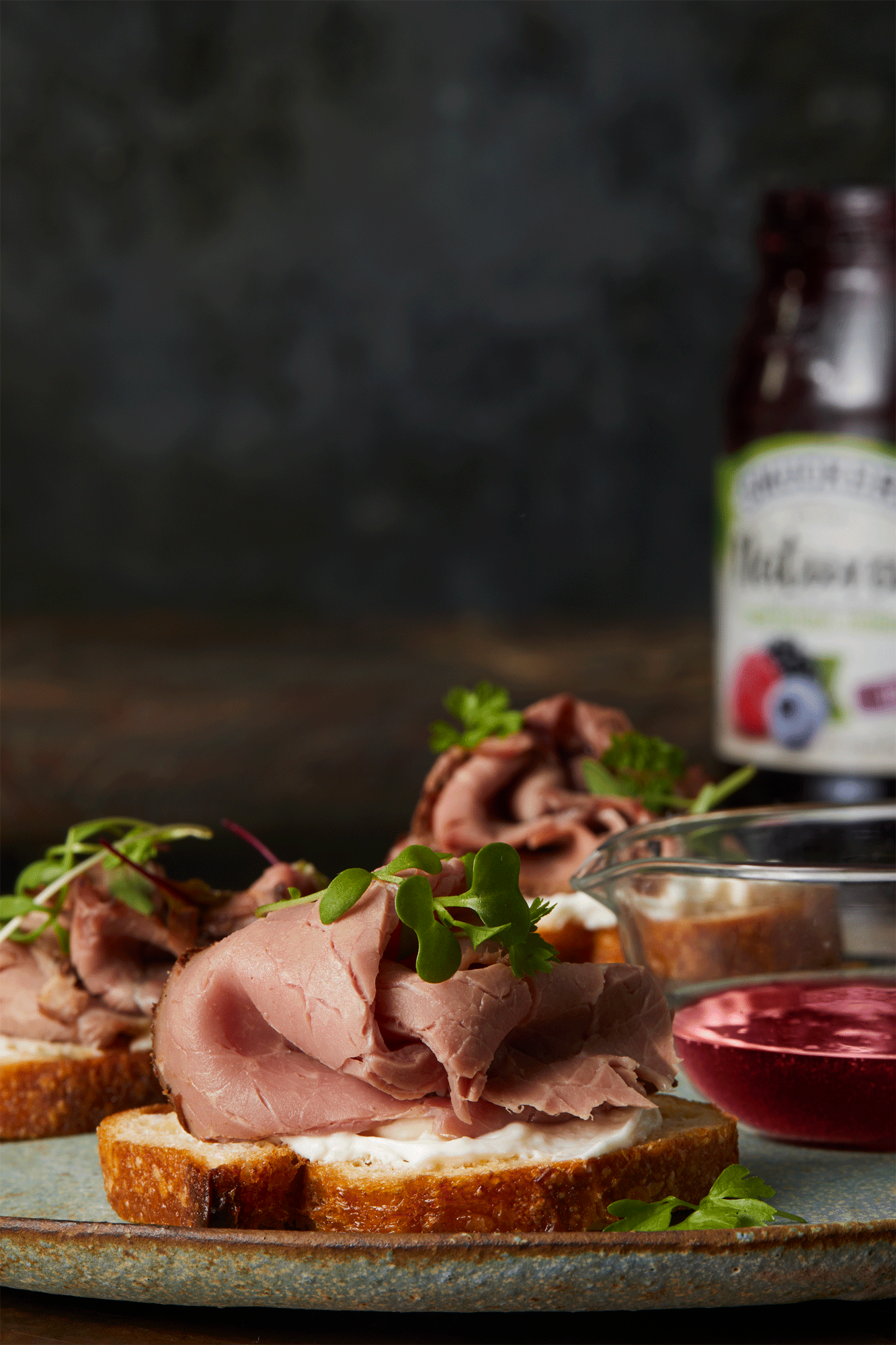 Close-up of sliced roast beef open-faced sandwiches with sprouts on a plate, accompanied by a jar of berry preserves and dipping sauces.