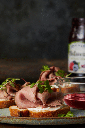 Close-up of sliced roast beef open-faced sandwiches with sprouts on a plate, accompanied by a jar of berry preserves and dipping sauces.