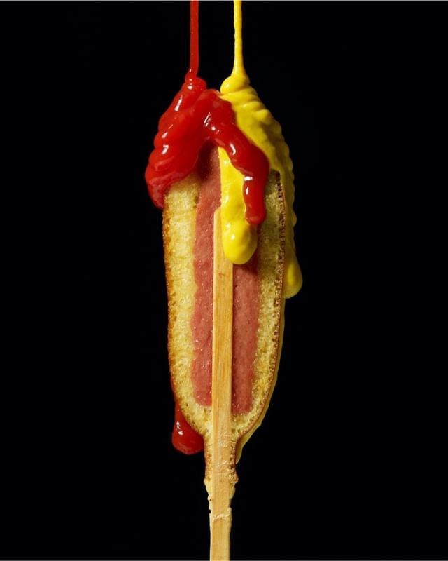 Corn dog on a stick with dripping ketchup and mustard against a black backdrop, showcasing food photography and culinary presentation.