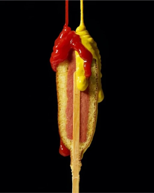 Corn dog on a stick with dripping ketchup and mustard against a black backdrop, showcasing food photography and culinary presentation.