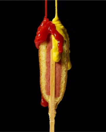 Corn dog on a stick with dripping ketchup and mustard against a black backdrop, showcasing food photography and culinary presentation.