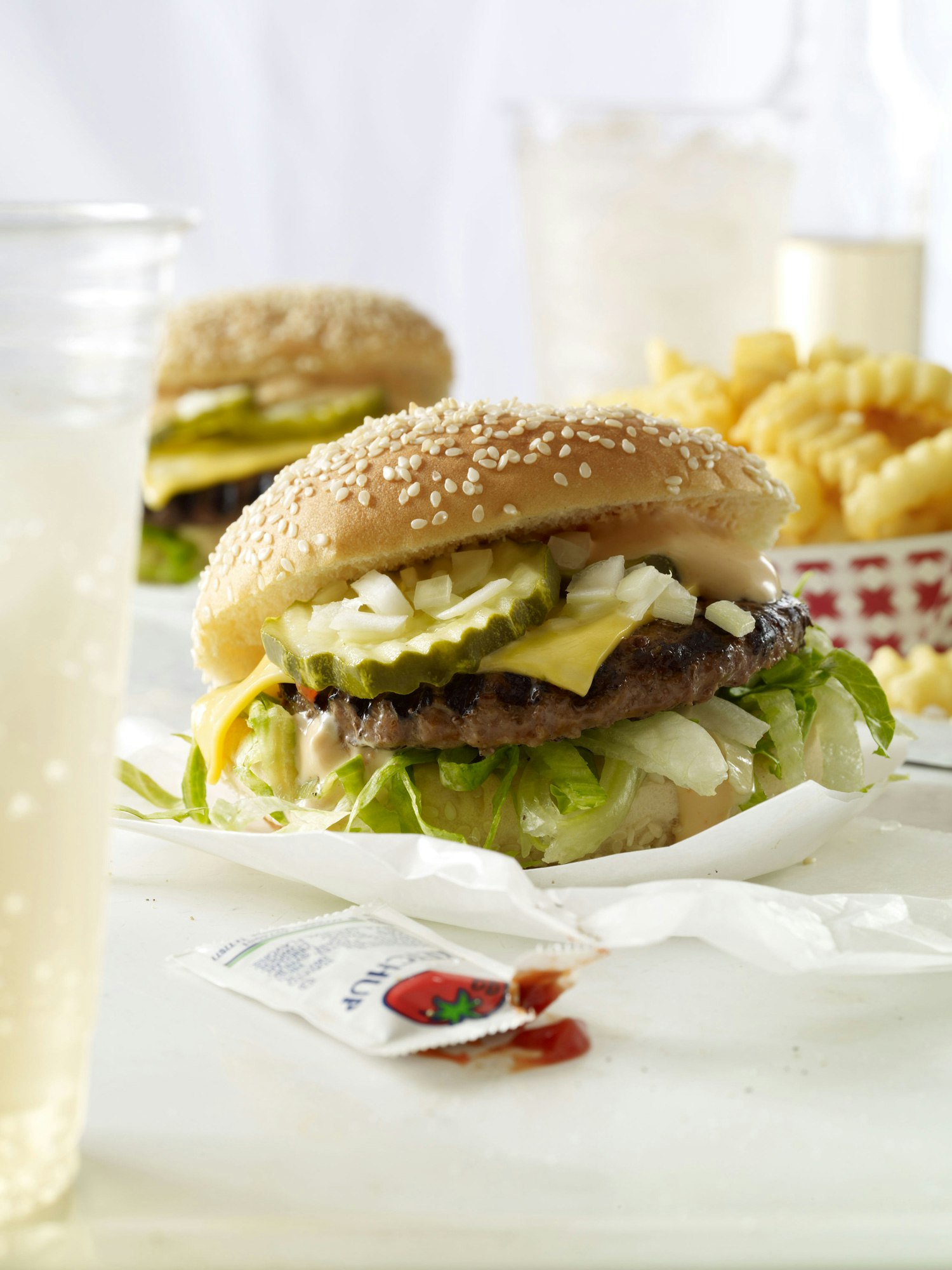 Close-up shot of a hamburger with sesame seed bun, lettuce, cheese, pickles, onions, and sauce, served with fries and a drink in a white setting.