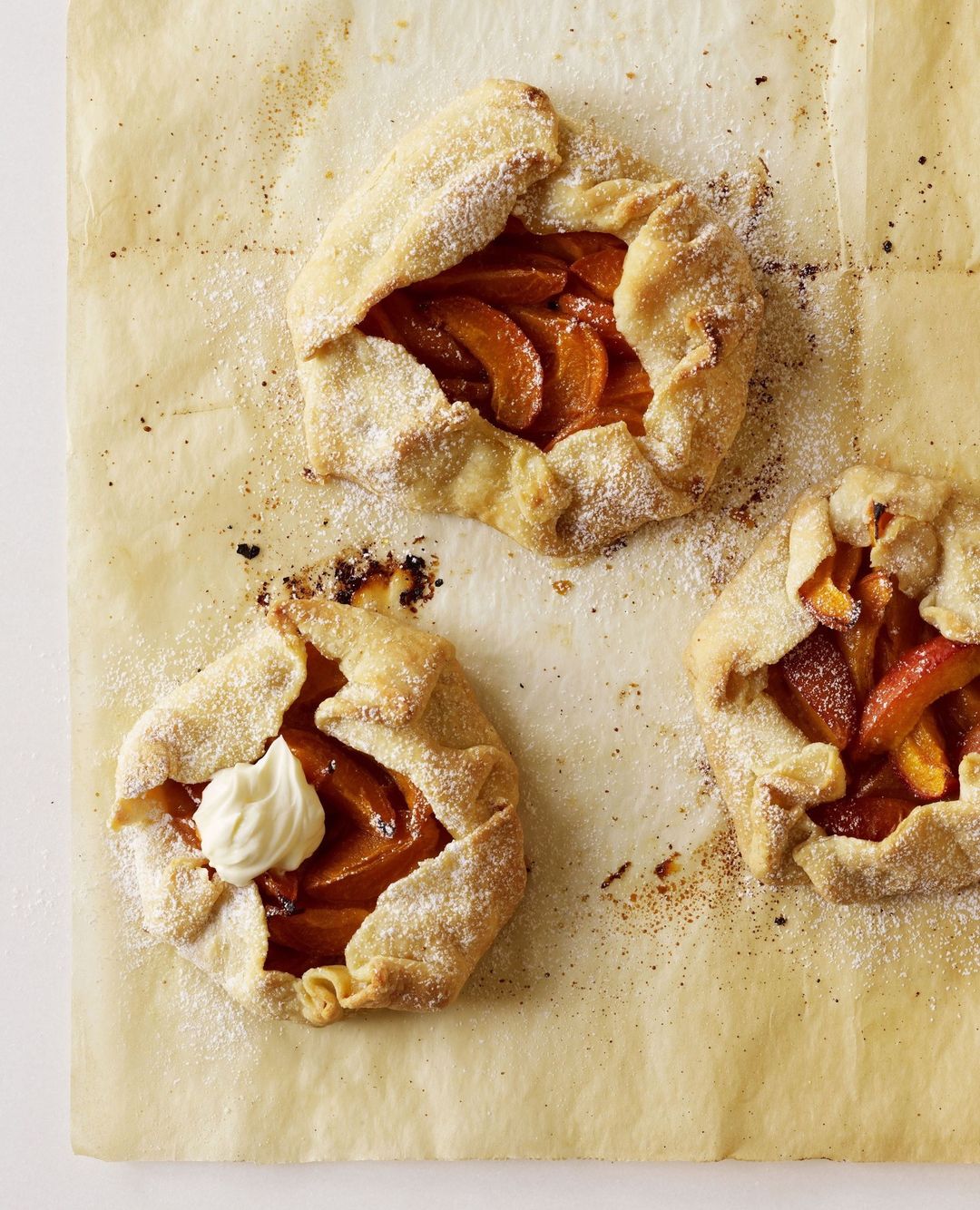 Overhead close-up of three rustic peach galettes dusted with powdered sugar, displayed on parchment paper for a delightful food still life.