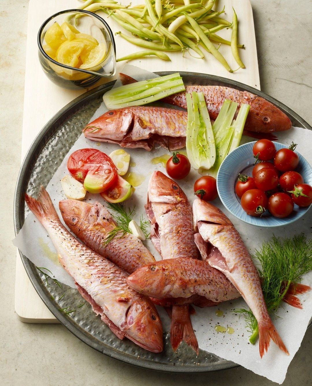Overhead shot of fresh red mullet fish on a metal tray with tomatoes, beans, cucumbers, herbs, and lemon slices ready for cooking.