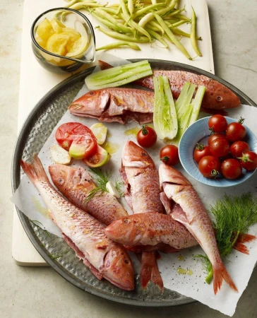 Overhead shot of fresh red mullet fish on a metal tray with tomatoes, beans, cucumbers, herbs, and lemon slices ready for cooking.