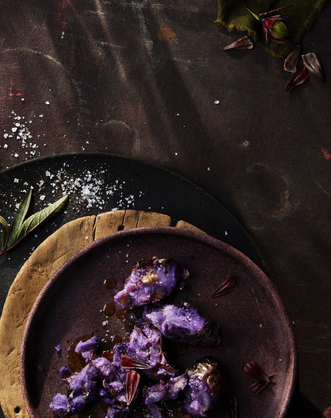Overhead shot of cooked purple sweet potatoes with seasoning and herb garnish on a purple plate, atop wood and slate surfaces.