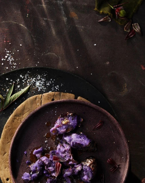 Overhead shot of cooked purple sweet potatoes with seasoning and herb garnish on a purple plate, atop wood and slate surfaces.