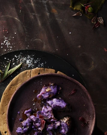 Overhead shot of cooked purple sweet potatoes with seasoning and herb garnish on a purple plate, atop wood and slate surfaces.