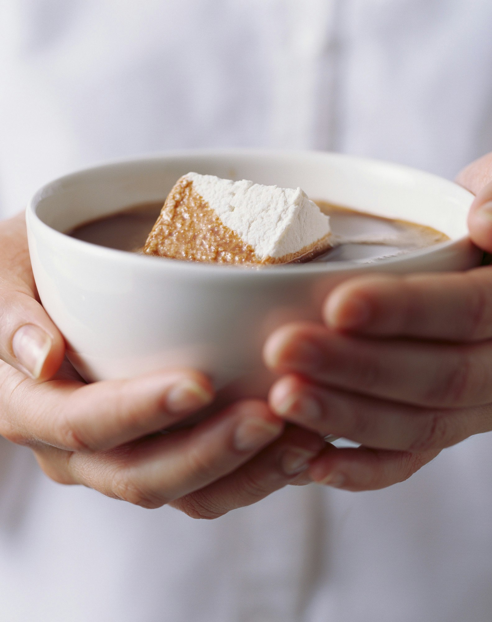 Close-up of a person holding a white bowl filled with hot chocolate and a large marshmallow, showcasing a sweet treat.