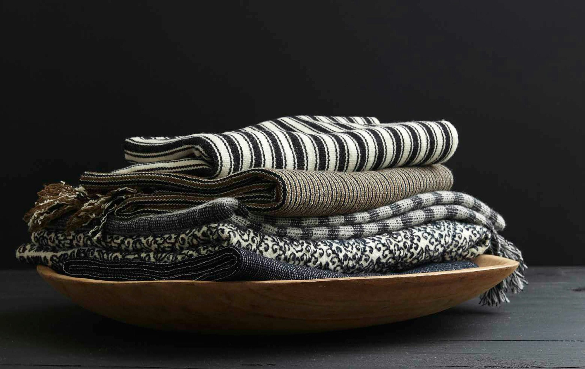 Stack of folded textile throws in a wooden bowl, against a black background. Includes striped, textured, and patterned textiles.