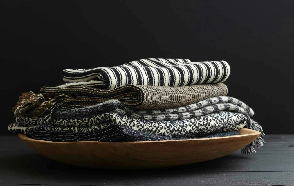 Stack of folded textile throws in a wooden bowl, against a black background. Includes striped, textured, and patterned textiles.