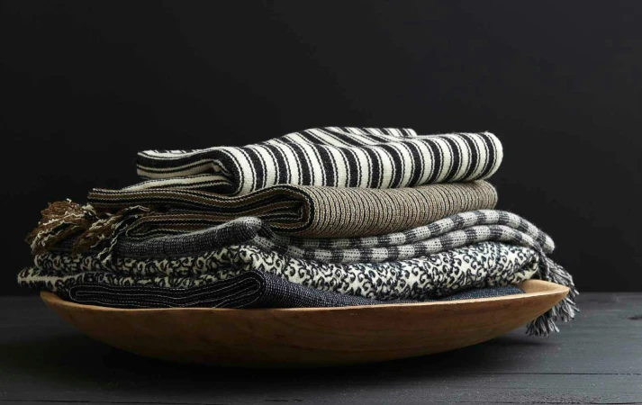 Stack of folded textile throws in a wooden bowl, against a black background. Includes striped, textured, and patterned textiles.