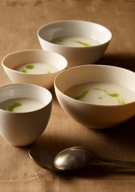 Four bowls of white soup with a swirl of green garnish, arranged on a tan linen surface with two silver spoons.