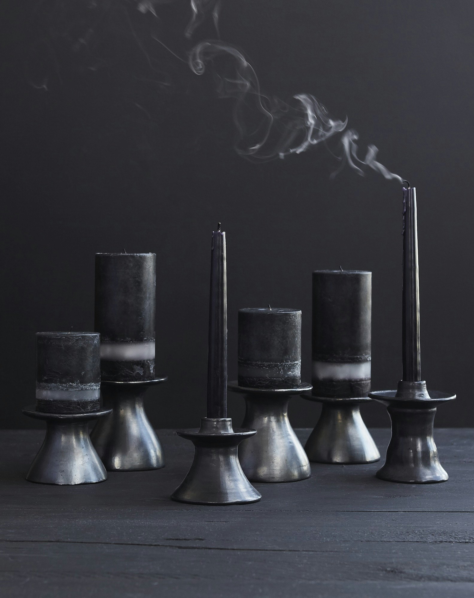 Still life of extinguished black candles in silver candle holders on a dark wooden surface with smoke rising in a dim setting.
