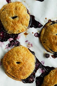 Overhead shot of golden blueberry hand pies on parchment paper with blueberry filling spilling out, creating a rustic and delicious food scene.