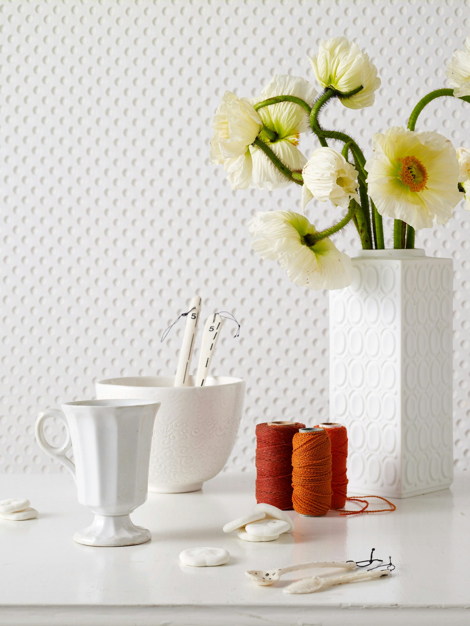 White flowers in a patterned vase, alongside spools of thread, a bowl, cup, crafting tools and embellishments arranged on a white surface.