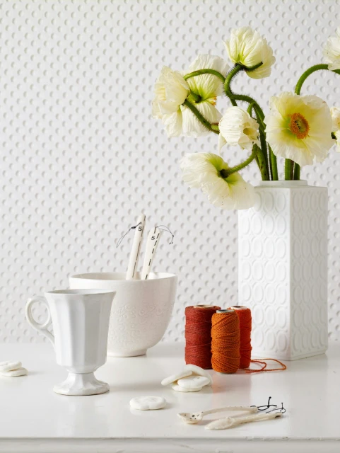White flowers in a patterned vase, alongside spools of thread, a bowl, cup, crafting tools and embellishments arranged on a white surface.
