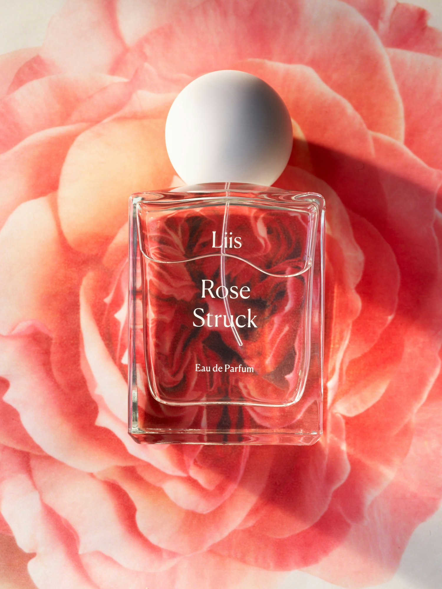 Rectangular perfume bottle with a white spherical cap, featuring the brand name and product on a soft-focus pink rose backdrop.