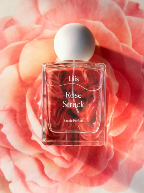 Rectangular perfume bottle with a white spherical cap, featuring the brand name and product on a soft-focus pink rose backdrop.