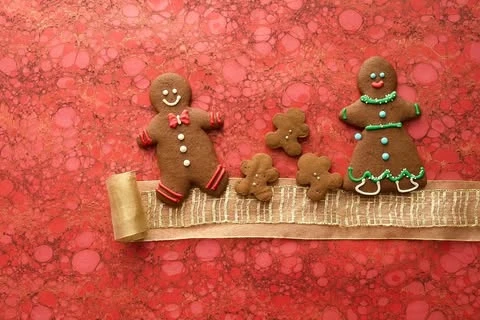 Gingerbread cookies arranged on a decorative ribbon against a textured red background, featuring a gingerbread man and woman.