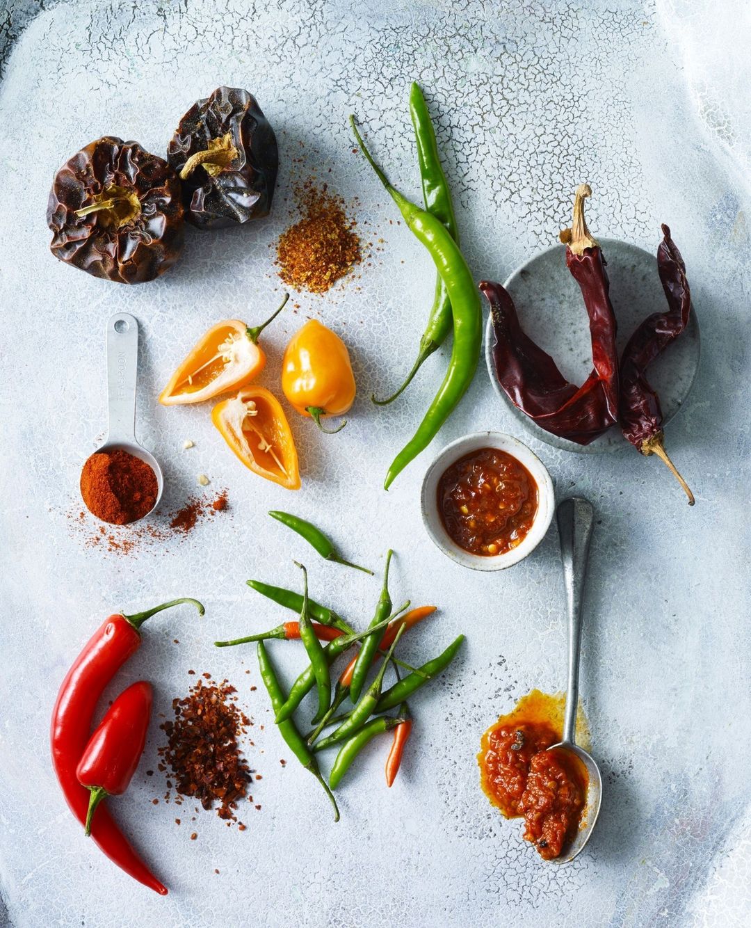 Overhead view of red, green, and yellow chili peppers, dried chilies, powders and pastes, and measuring spoons on a white speckled surface.