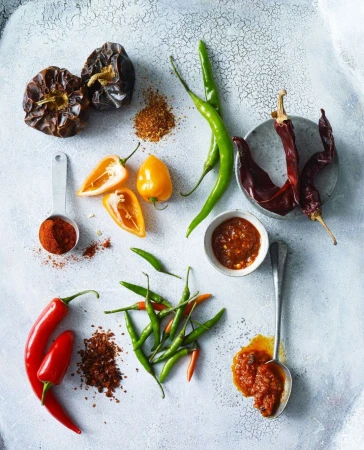 Overhead view of red, green, and yellow chili peppers, dried chilies, powders and pastes, and measuring spoons on a white speckled surface.