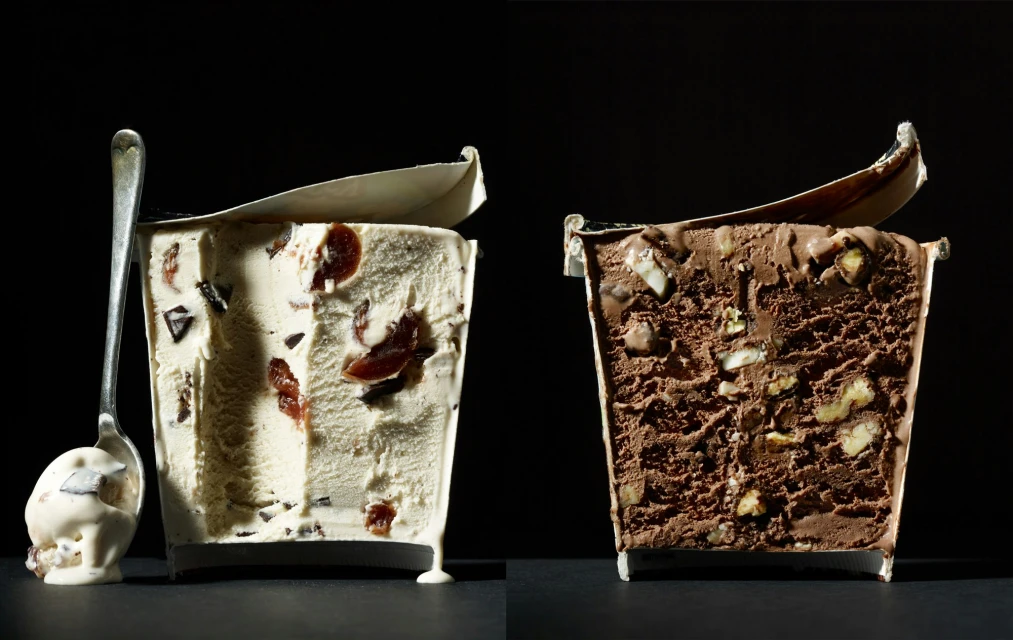 Two containers of ice cream, chocolate with nuts and vanilla with cherries, with a spoon scooping vanilla ice cream.