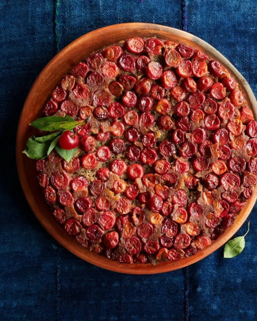 Overhead shot of a cherry cake on a wooden plate, adorned with fresh cherries and leaves against a dark blue fabric backdrop.