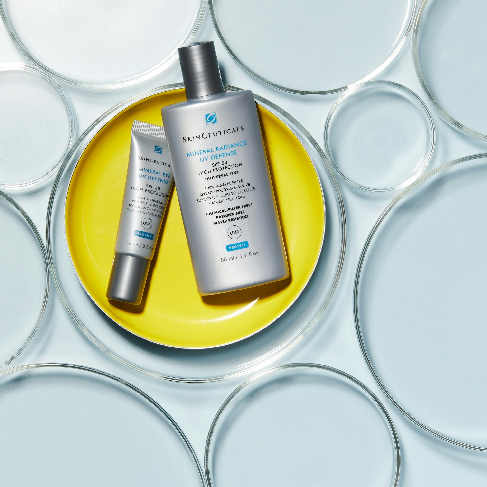 Two bottles of SkinCeuticals sunscreen on a yellow plate, surrounded by petri dishes on a light blue background.