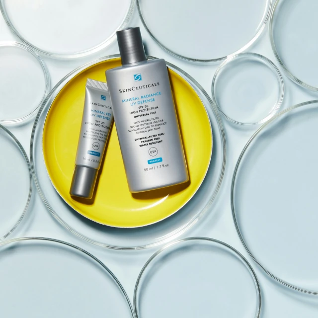 Two bottles of SkinCeuticals sunscreen on a yellow plate, surrounded by petri dishes on a light blue background.
