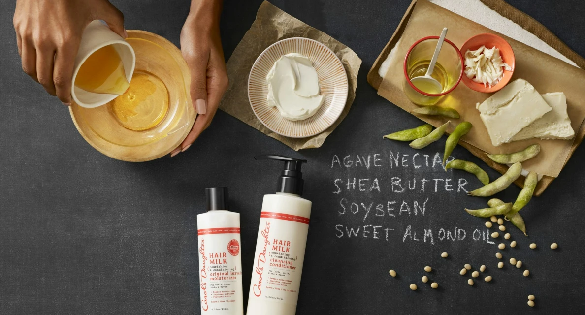 Overhead shot: person pours liquid into bowl. Products, agave, shea butter, soybean, almond oil, and ingredients on a dark surface.