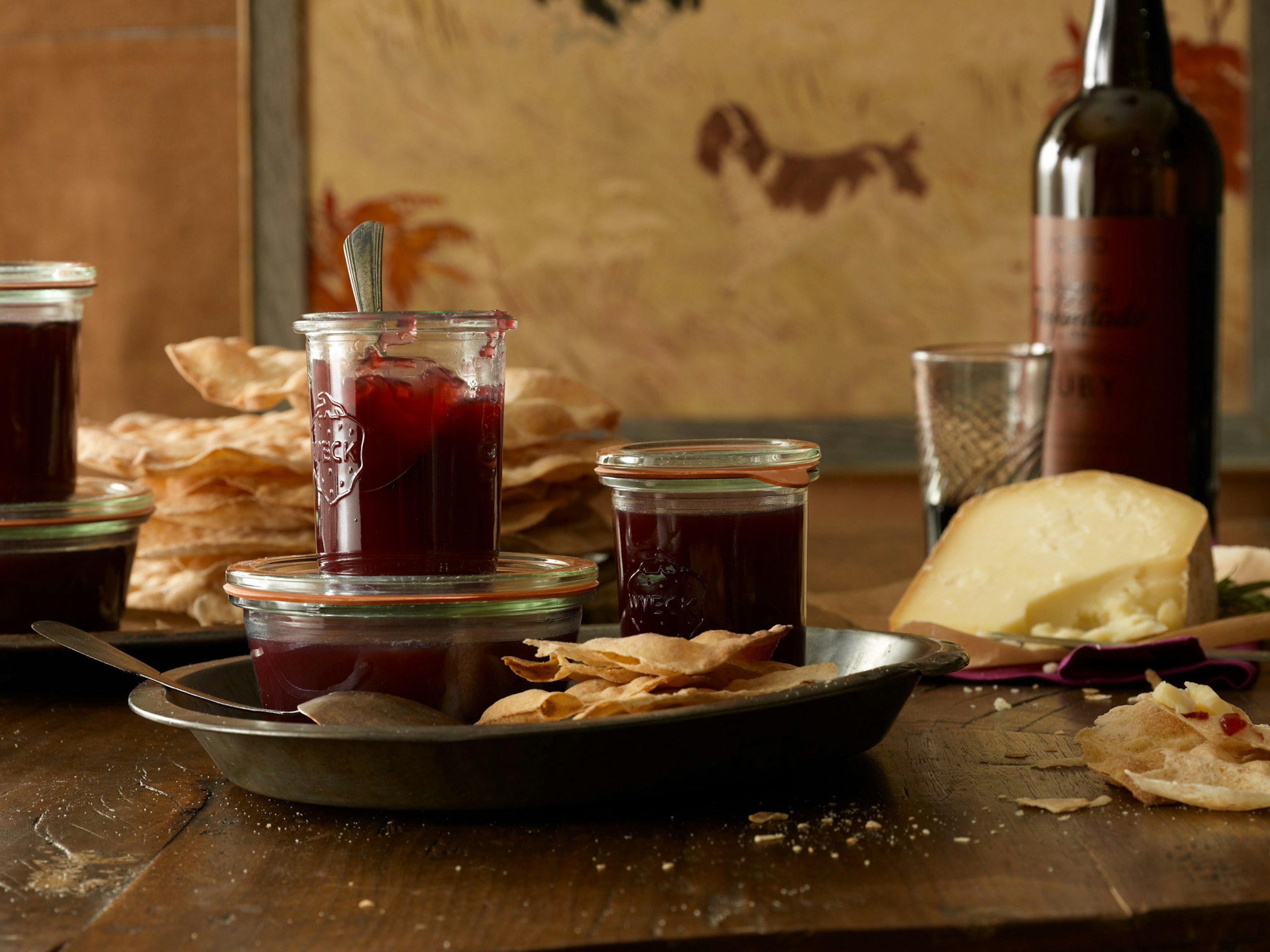 Jars of plum jam, cream colored crackers, and a wedge of cheese sit on a wooden table in a naturally lit indoor setting.