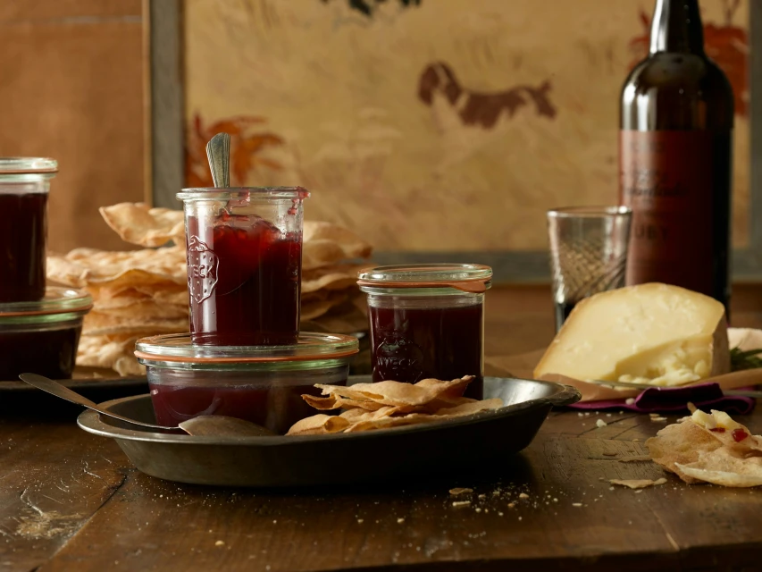 Jars of plum jam, cream colored crackers, and a wedge of cheese sit on a wooden table in a naturally lit indoor setting.