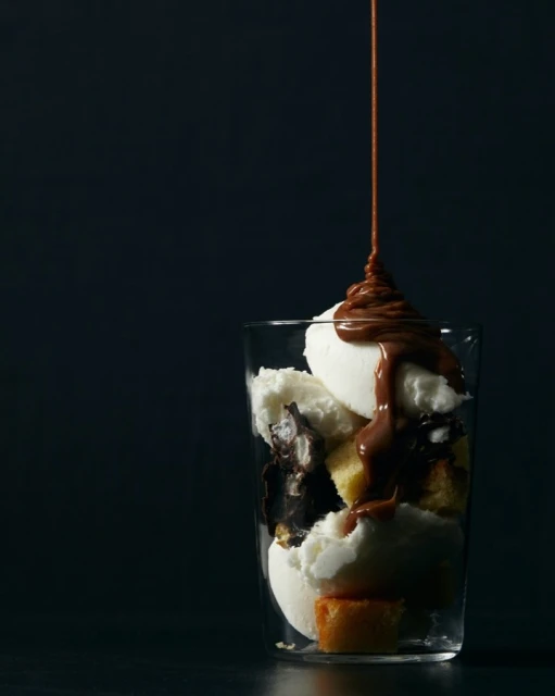 Ice cream sundae with chocolate syrup drizzled in a glass against a dark background. Dessert includes cake and chocolate pieces.
