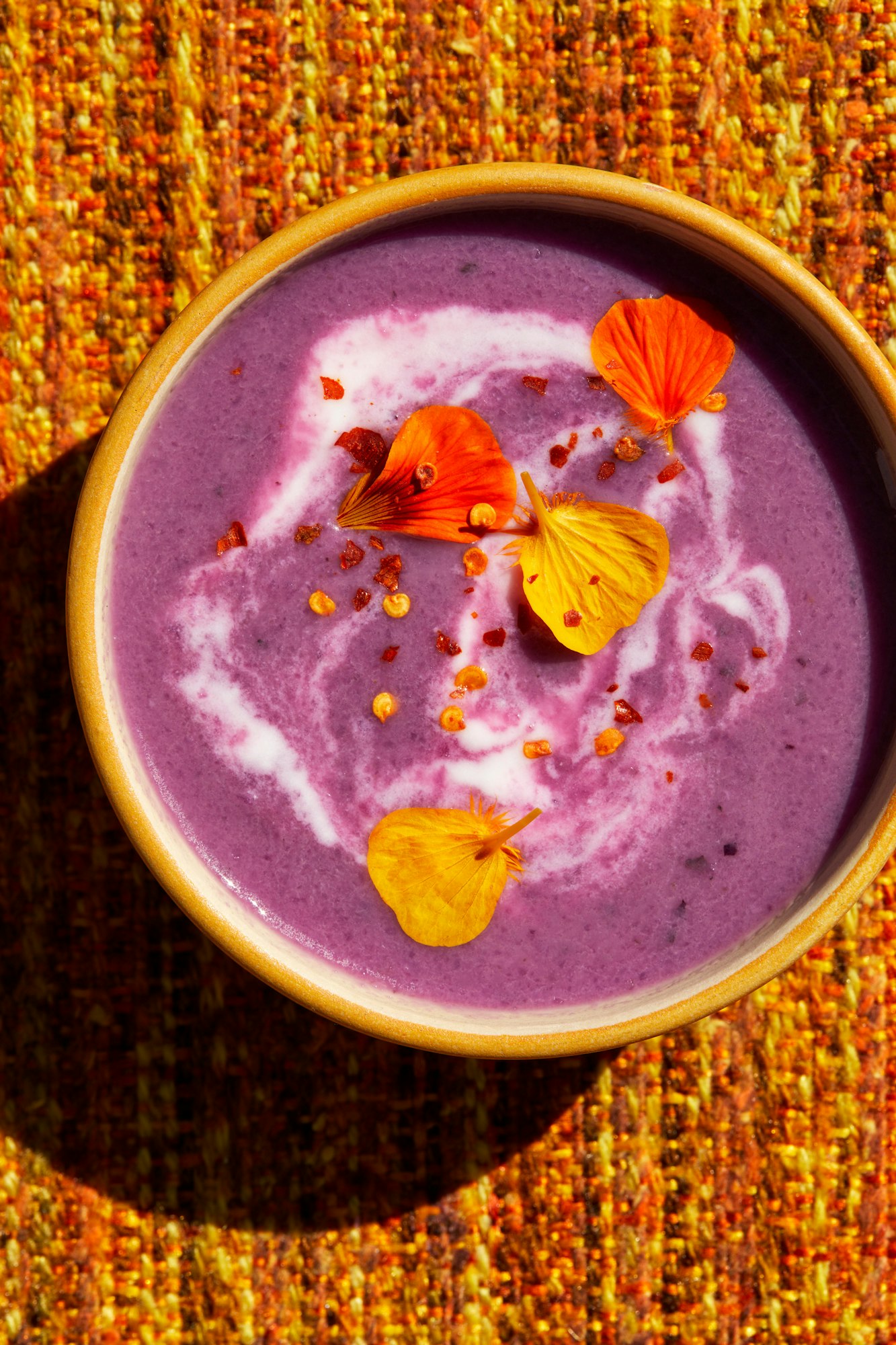 Overhead shot of a bowl of purple soup with a swirl of white cream, garnished with edible flowers, on a textured orange fabric.