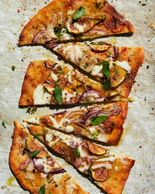 Overhead view of sliced homemade pizza with figs, red onion, fresh herbs, and cheese on parchment paper, food photography.