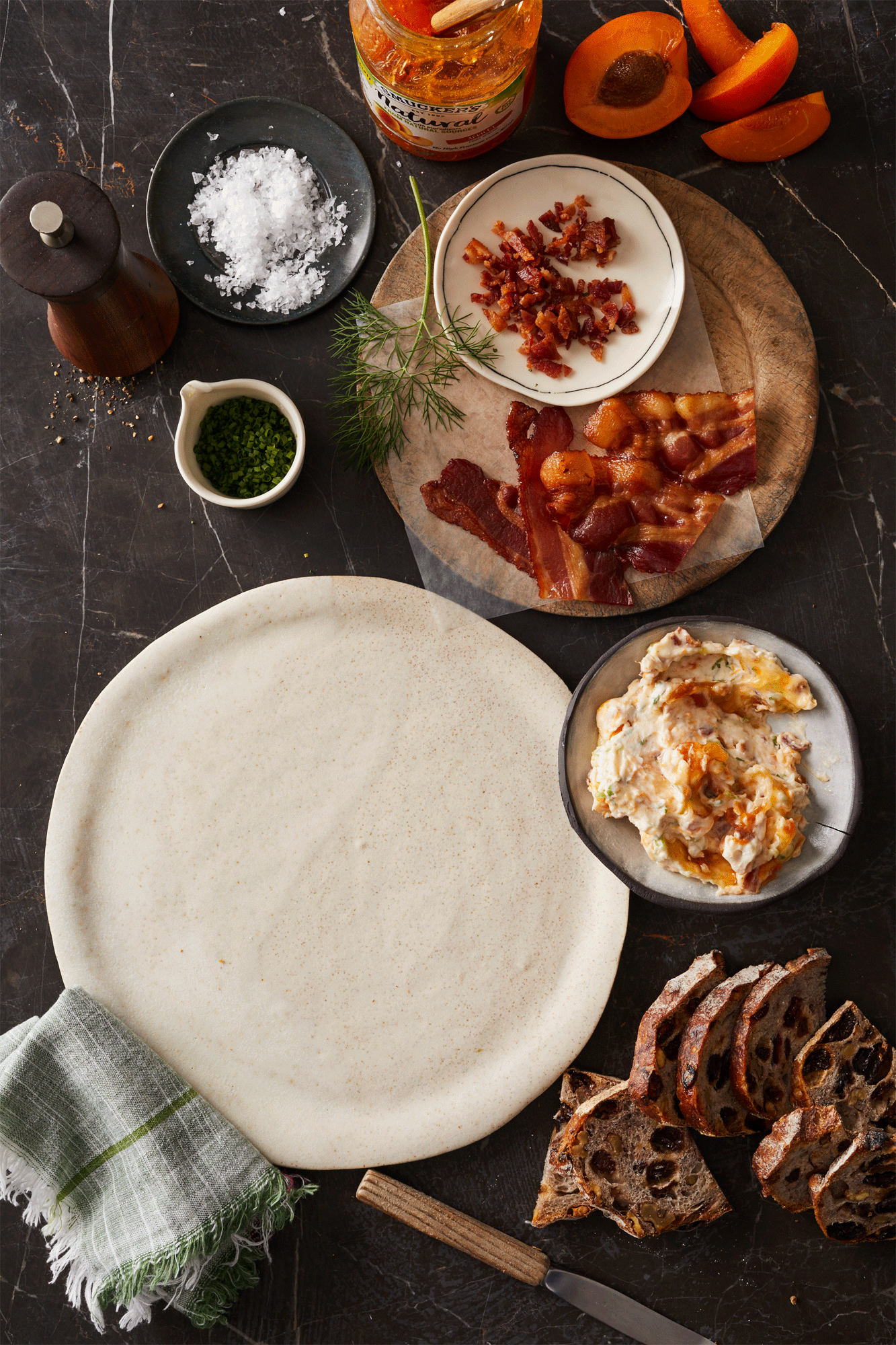 Overhead shot of a rustic table laden with a charcuterie board, sliced bread, bacon jam, apricots, herbs, and a serving plate.