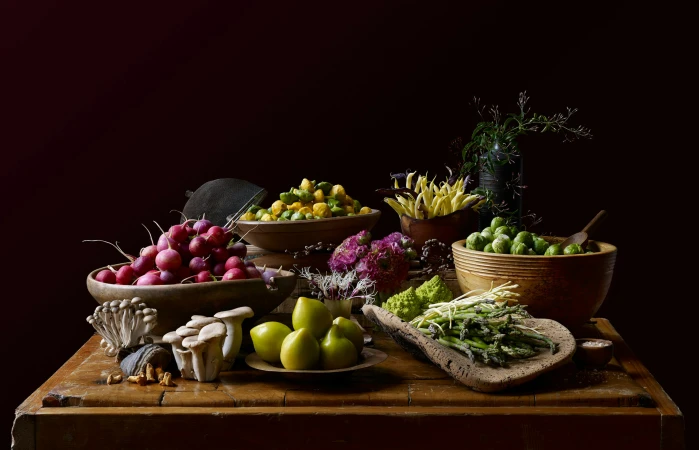 A still life featuring an arrangement of radishes, brussel sprouts, mushrooms, plums, asparagus, and flowers on a wooden table.