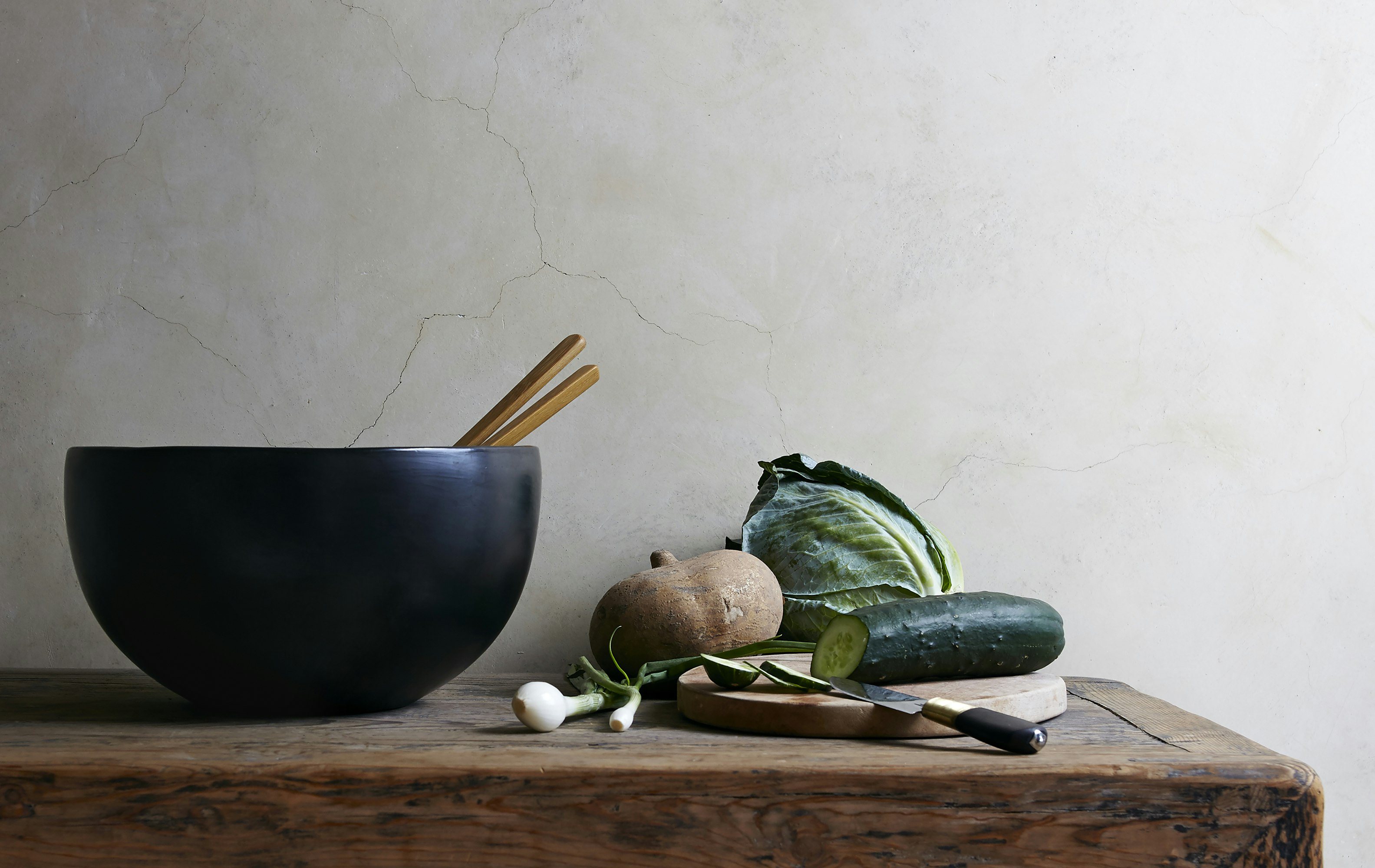 Still life captures a black bowl filled with chopsticks, accompanied by various vegetables on a rustic wooden table against a neutral backdrop.