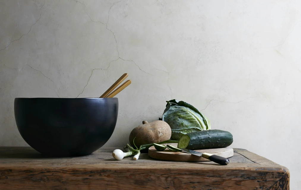 Still life captures a black bowl filled with chopsticks, accompanied by various vegetables on a rustic wooden table against a neutral backdrop.