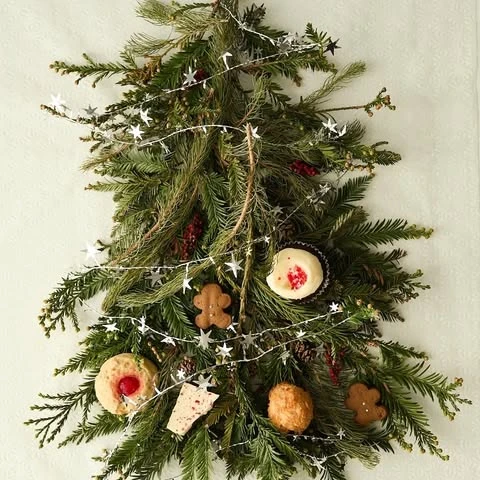 Holiday dessert arrangement featuring an evergreen branch decorated with star garland and festive cookies and sweets on a white backdrop.