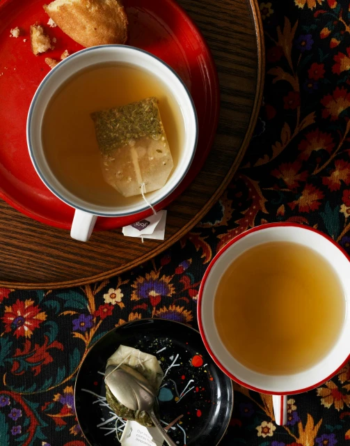 Overhead shot of cups of tea with teabags, small black dish with used teabag and spoon, and a plate of pastry on a patterned table.