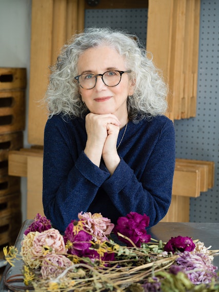 Portrait of a mature woman with gray curly hair, wearing glasses and a navy sweater, with dried flowers on a metal table.