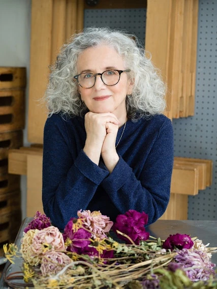 Portrait of a mature woman with gray curly hair, wearing glasses and a navy sweater, with dried flowers on a metal table.