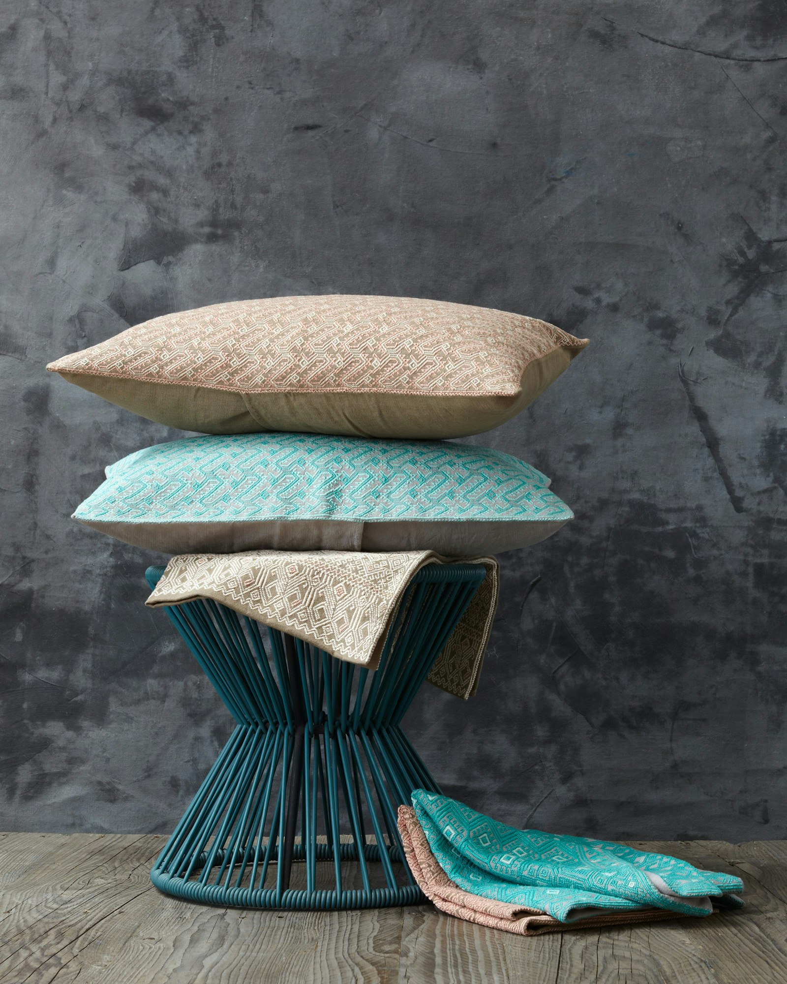 Still life of stacked decorative pillows and folded textiles on a woven stool against a textured gray wall and wood floor.