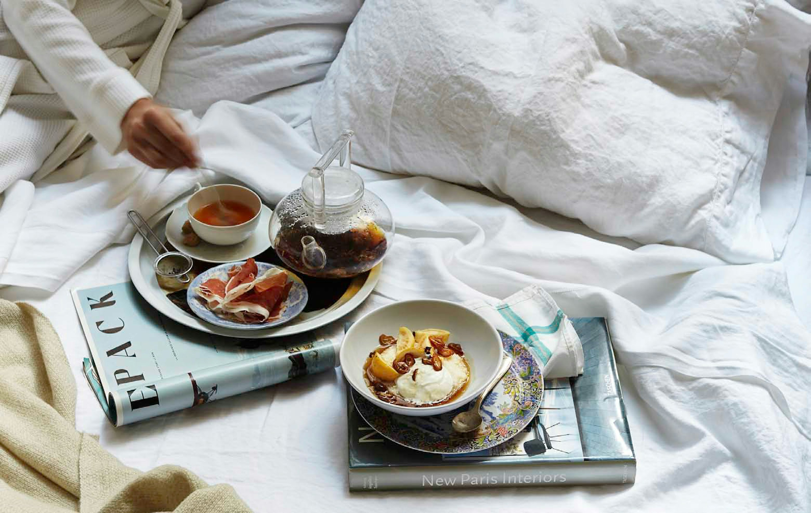 Overhead shot of a served tea and appetizer breakfast in bed featuring a teapot, teacup, and a plate of cured meat.