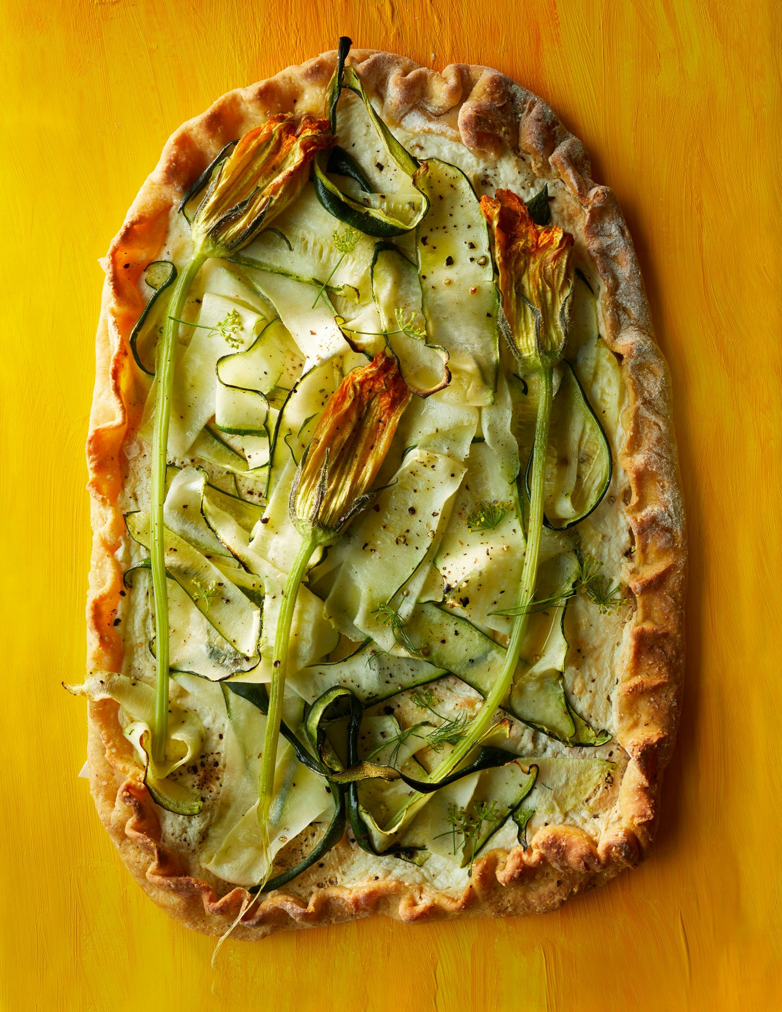 Overhead shot of a zucchini and squash blossom tart on a rustic crust, featuring spiral zucchini slices and yellow edible flowers on a yellow background.