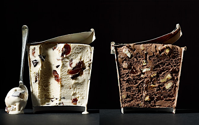 Two containers of ice cream side-by-side, one vanilla with cherries and chocolate chunks and the other chocolate with nuts, with a spoon.