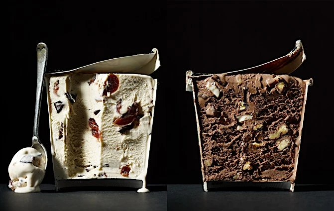 Two containers of ice cream side-by-side, one vanilla with cherries and chocolate chunks and the other chocolate with nuts, with a spoon.